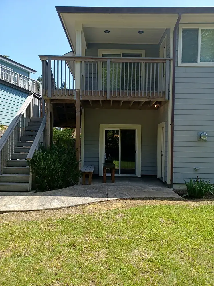 A two-story house in Shelter Cove with a light grey exterior and a wooden staircase on the left leading to a balcony on the upper level. The lower level features a glass door flanked by windows, and a small patio area with a concrete walkway in front of the house, bordered by a grassy lawn.