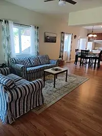 A cozy living room in Shelter Cove with a striped sofa and armchair, a coffee table, and a patterned rug on wooden flooring. A dining area with a table and chairs is visible in the background, along with a kitchen. Sunlight streams through large windows with patterned curtains.