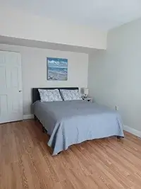 A tidy bedroom with a wooden floor features a bed with a gray bedspread and two pillows. Above the bed is an ocean-themed painting reminiscent of Shelter Cove. To the right of the bed is a bedside table with a lamp. A closed white door is visible on the left.