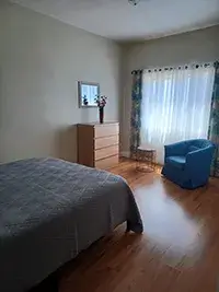 A bedroom in Shelter Cove with a wooden floor features a neatly made bed with a gray comforter, a wooden dresser with a small mirror above it, and a blue armchair next to sheer, light-filtering curtains. The room is brightly lit by natural light from the large window.