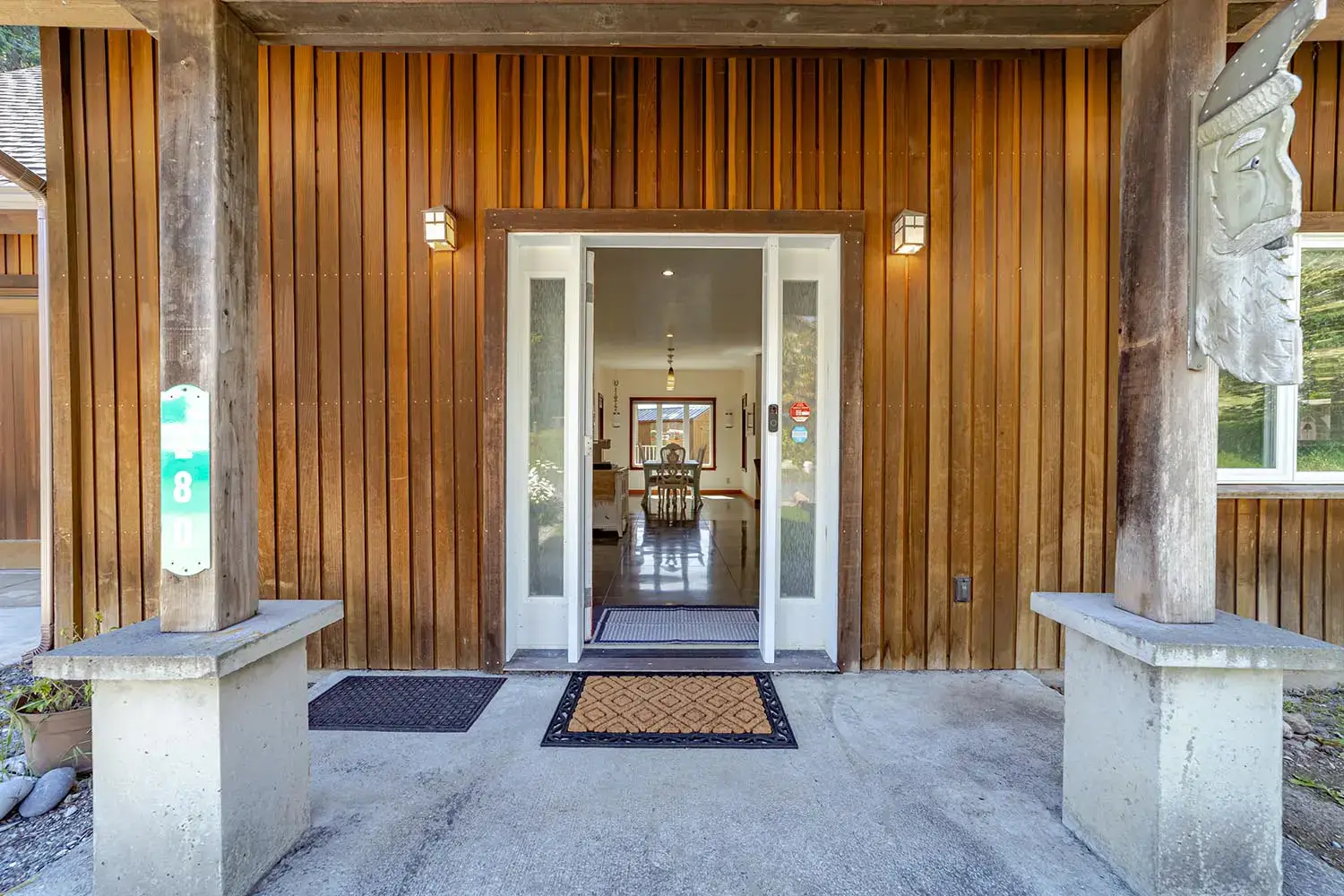 The front entrance of this charming Shelter Cove wooden house features vertical paneling and a glass-paneled door flanked by narrow windows. Two doormats adorn the concrete porch, supported by wooden pillars. Inside, a chandelier illuminates the dining area, making it an inviting Air B&B retreat.