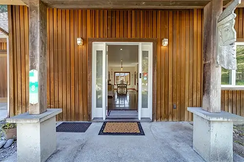The front entrance of this charming Shelter Cove wooden house features vertical paneling and a glass-paneled door flanked by narrow windows. Two doormats adorn the concrete porch, supported by wooden pillars. Inside, a chandelier illuminates the dining area, making it an inviting Air B&B retreat.