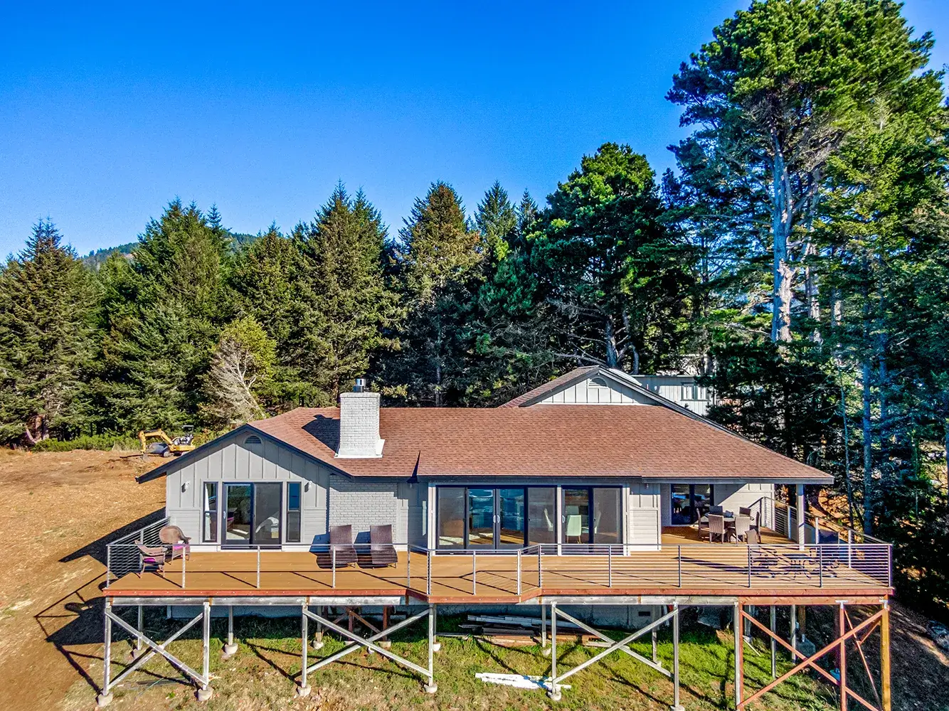 A house with a large wooden deck on stilts, nestled in Shelter Cove and surrounded by lush green trees. The house has a brown roof and several large windows. Outdoor furniture is arranged on the deck, which overlooks a grassy area. The sky is clear and blue, suggesting a sunny day.