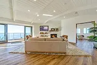 A spacious living room with a high, white-paneled ceiling and large windows offering an ocean view of Shelter Cove. It features a beige sectional sofa, a fireplace with a mounted TV above, wooden flooring, and a door leading to another room. A potted plant decorates the corner.