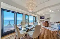 A modern dining room with a glass table and six white chairs, adjacent to a living area with a couch, armchair, and fireplace. Large windows offer a panoramic ocean view of Shelter Cove. A chandelier hangs from the ceiling, and the hardwood floor adds warmth to the space.