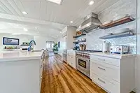 A modern kitchen with wooden floors and white cabinetry. It features a large island with a sink, a stainless steel oven and range hood, open shelving, a backsplash of white tiles, and recessed lighting in the ceiling—truly resembling the elegance of Shelter Cove.