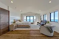 Modern bedroom with large windows showcasing a scenic ocean view at Shelter Cove. The room features a wooden floor, a cozy bed with white linens, a stylish armchair, a nightstand, and a balcony with glass doors. Bright, minimalist decor enhances the airy atmosphere.