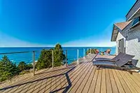A spacious wooden deck overlooking a calm ocean at Shelter Cove, extending from the side of a modern house. The deck is equipped with lounge chairs and bordered by transparent railings. In the distance, the clear blue sky meets the serene waters. Trees are partially visible below the deck.