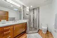 A modern bathroom in Shelter Cove with a wooden vanity featuring dual sinks and a large mirror. To the right is a glass-enclosed shower with tiled walls and a white toilet. The floor is wooden, and the overall design is minimalist with clean lines and neutral colors.