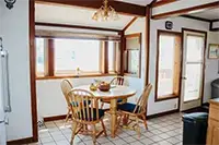 A cozy dining area with a wooden round table and four matching chairs overlooks the natural beauty of Shelter Cove. The table is set near large windows that allow light to fill the room, while a chandelier hangs above. A door to the right opens up to more stunning views. The floor is tiled.