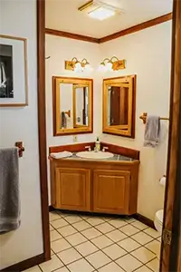 A bathroom at Shelter Cove features a wooden double vanity with two rectangular mirrors and wall-mounted light fixtures above each mirror. The space has white walls, tiled flooring, and towels hanging on racks.