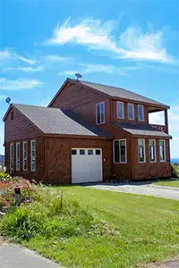 A two-story wooden house with a sloped gray roof and large windows sits elegantly in Shelter Cove. The house boasts a garage and is surrounded by a well-maintained lawn and a flower bed on the left. The sky above is bright blue with wispy clouds.