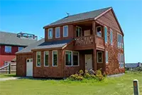 A two-story house with a wooden exterior is set against a blue sky in Shelter Cove. The house features multiple windows, a small balcony on the upper level, and a lush green lawn with some shrubs in the front yard. Another house is visible in the background.