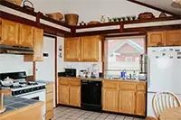 A cozy kitchen with wooden cabinets, a gas stove, and a white refrigerator adorned with magnets. A window above the sink lets in natural light, highlighting a counter with various kitchen appliances and utensils. In the foreground, a small dining table and chairs evoke the charm of Shelter Cove.