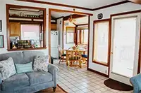 A cozy living area featuring a gray couch with patterned cushions. Beyond the couch is a kitchen with wooden cabinets and a dining area with a wooden table and chairs. The space in Shelter Cove has a bright atmosphere thanks to large windows and a glass-paneled door.