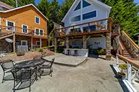 A spacious patio featuring a round metal table with four chairs in the foreground is set against the backdrop of two multi-story houses, one of wooden siding and the other with large windows and a modern design. Surrounded by lush greenery, it's an ideal spot for vacation rentals in Humboldt.