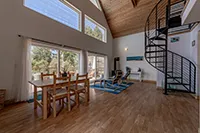 A spacious, modern living room with high ceilings, wooden floors, and large windows. A dining table with chairs is on the left and a sitting area with chairs and a rug on the right. A curved staircase leads to an upper level. Natural light fills the room in this prime Humboldt vacation rental.