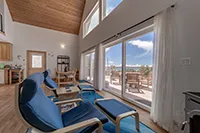 A bright, cozy living room with high ceilings and large windows awaits you in this Humboldt vacation rental. Blue chairs are arranged beside a coffee table on a blue rug. Glass doors lead to a sunny patio with a clear sky and ocean view. Light wooden floors and white walls enhance the airy feel.
