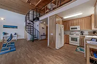 A compact kitchen with wooden cabinetry, a white refrigerator, oven, and microwave. Adjacent to the kitchen is a small dining area. A spiral staircase leads to a loft with metal railings. The space features wooden flooring and walls with muted colors—ideal for Humboldt vacation rentals.
