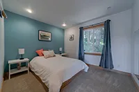 A cozy bedroom with a teal accent wall behind a neatly made bed with white bedding and pink pillows. The room, perfect for vacation rentals, has a large window with blue curtains, two bedside tables with lamps, and framed artwork on the walls. The floor is carpeted in light beige tones reminiscent of a Humboldt retreat.