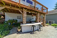 Outdoor patio area with stone columns supporting a wooden deck overhead. There is a concrete countertop with two stools, surrounded by potted plants. The shaded space underneath the deck offers a relaxing retreat, perfect for vacation rentals in Humboldt. A gray house with a window is visible in the background.
                  